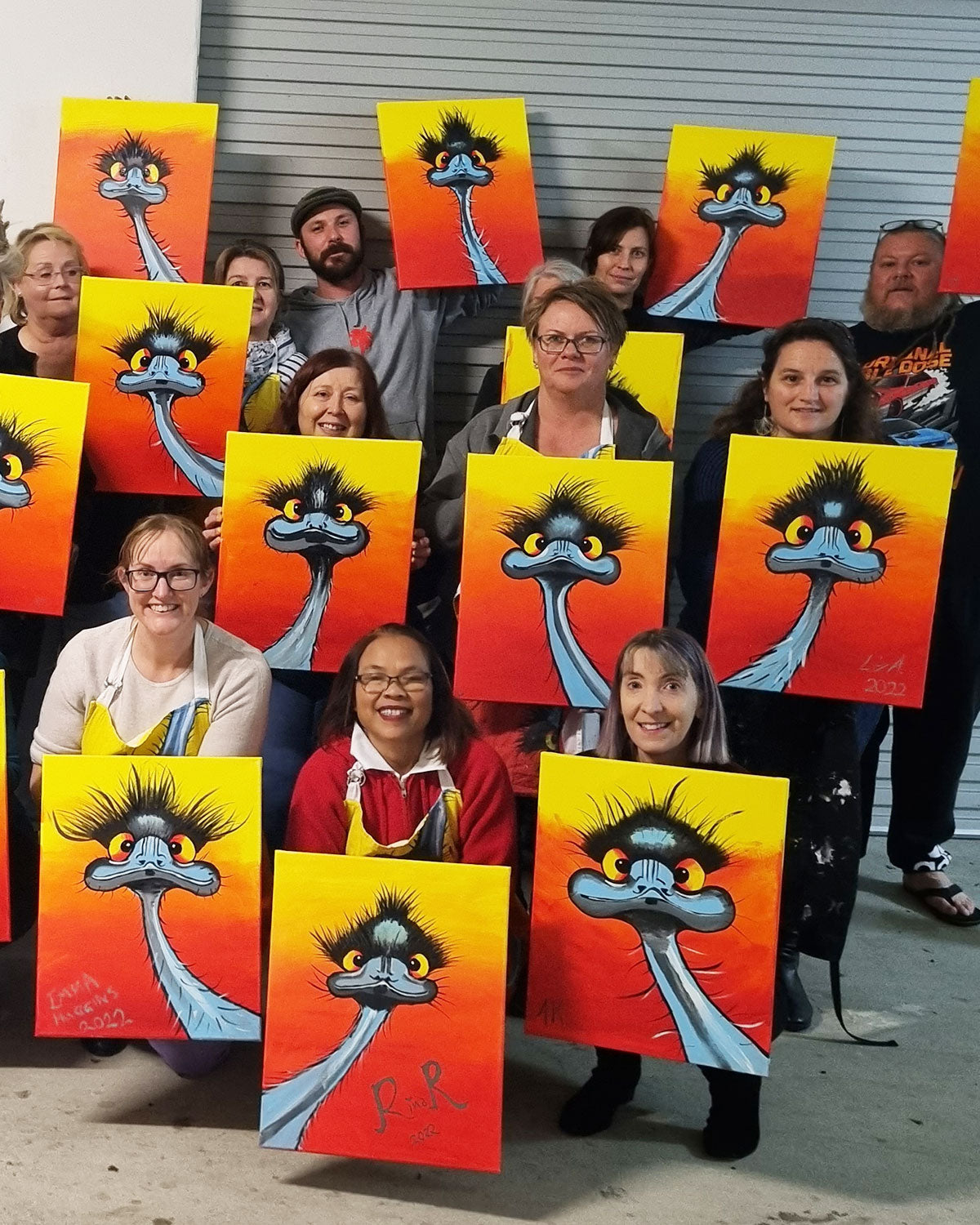 A group of people holding up hand-painted canvases featuring a cartoon emu character with a yellow and orange gradient background.