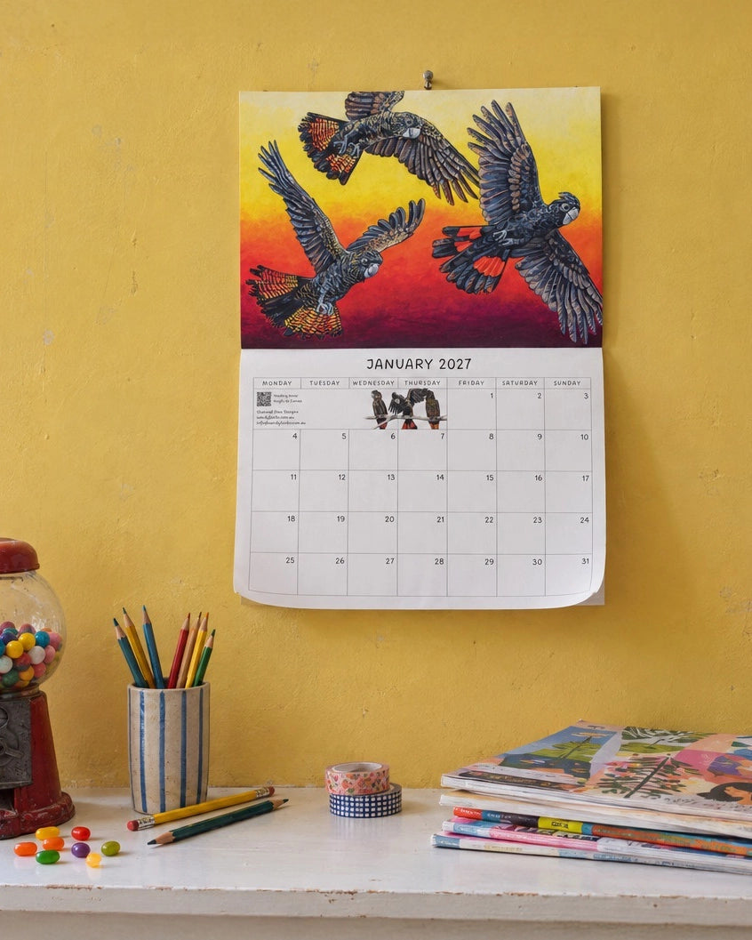 A January 2027 wall calendar featuring a colorful illustration of three flying cockatoos against a yellow and red sunset, hanging on a textured yellow wall above a desk with pencils, tape, and magazines.