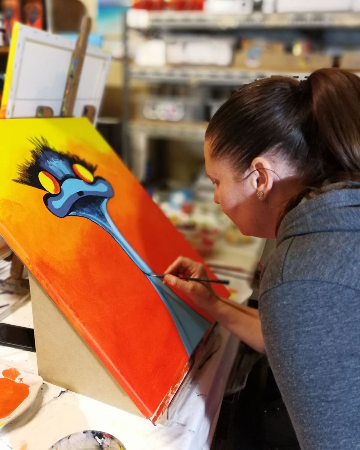 A woman painting a colorful canvas featuring a blue emu with large yellow eyes on a bright orange-yellow background, photographed in an art studio setting.