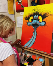 A woman painting a colorful portrait of an emu on a canvas set on an easel in an art studio.