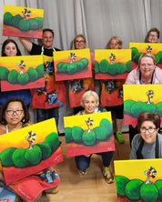 A group of people holding up colorful paintings of green emu eggs hatching on a bright yellow background with red shadows, set against a gray curtain backdrop.